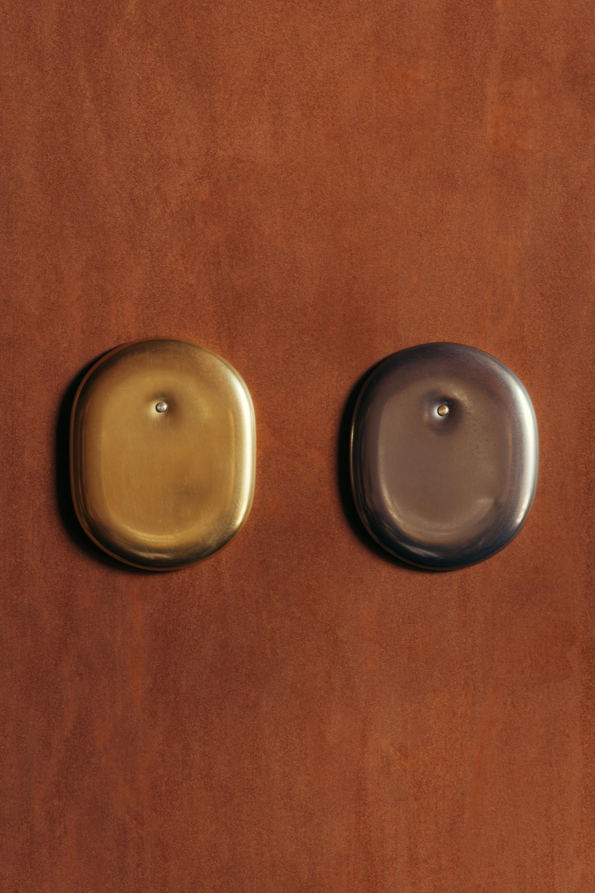  Another angle of the Andrea Walsh Small Wall Pieces against a brown plastered wall. On the left is the piece burnished in gold, and on the right, the piece burnished in white gold.