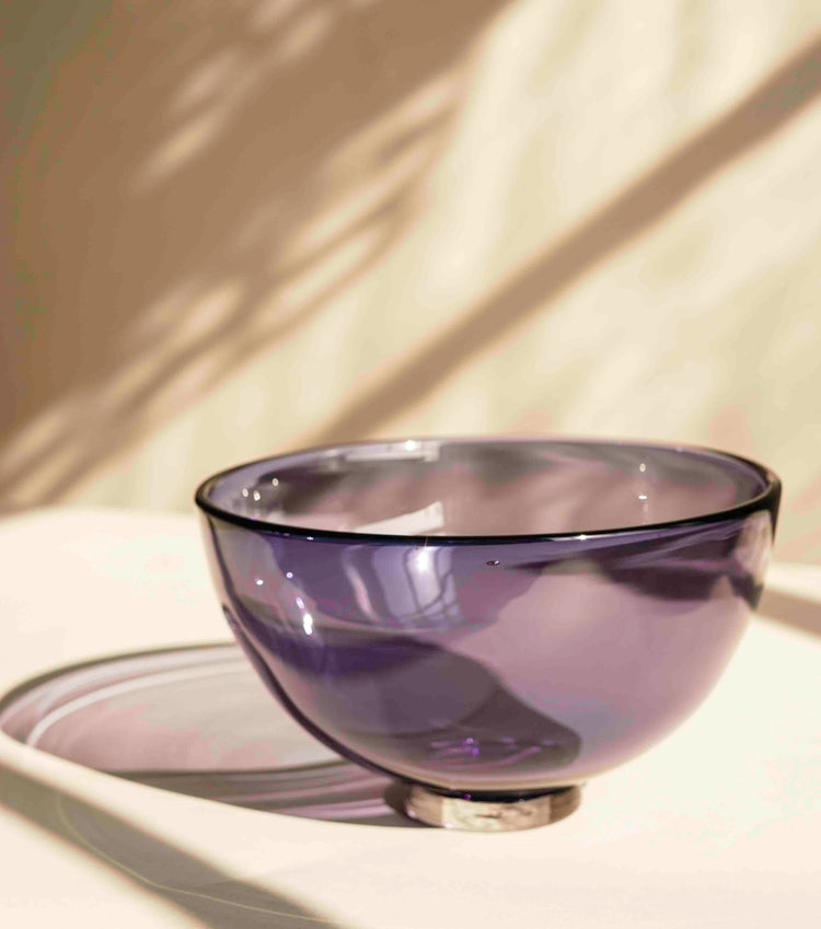 A violet purple glass bowl against a white background. A window on the right of the image casts shadows and light patterns on the bowl and the white backdrop behind.