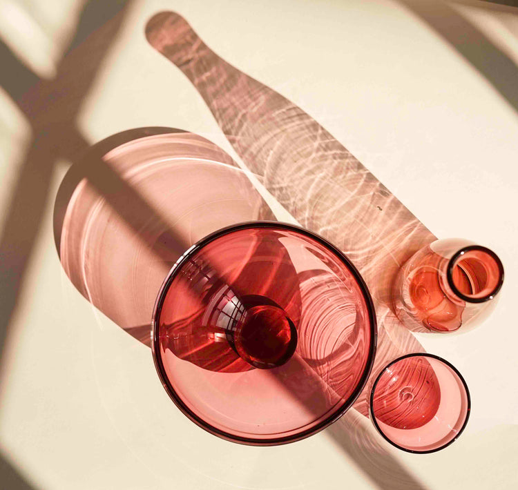 A collection of blush pink glassware from Lindean Mill, photographed from above on a white background. A gem bowl, a cup and a carafe are pictured.