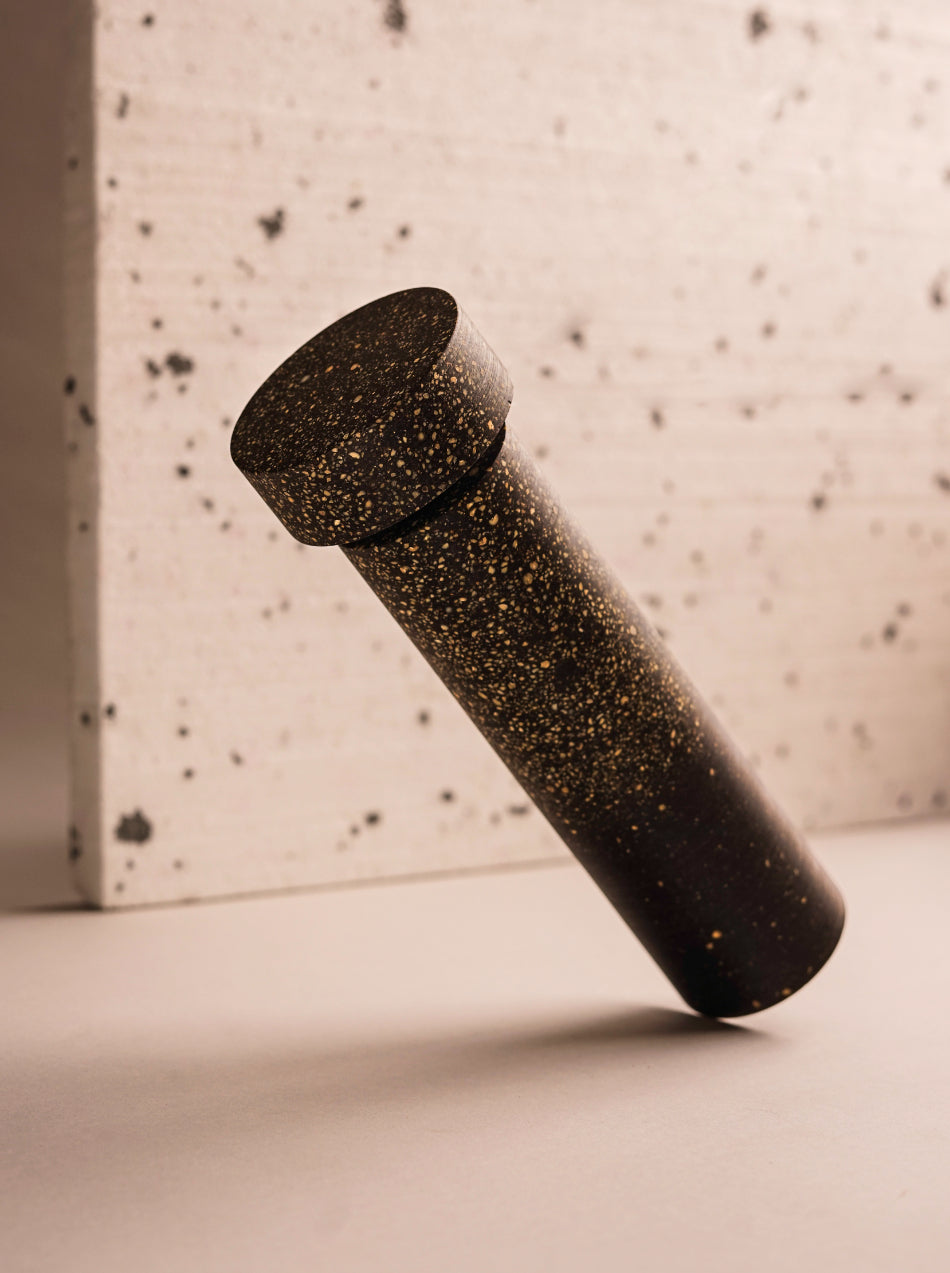  A pepper mill made from grey/brown bio resin and peppercorns. Lent to the left as if floating. In the background, a speckled piece of white packing foam.