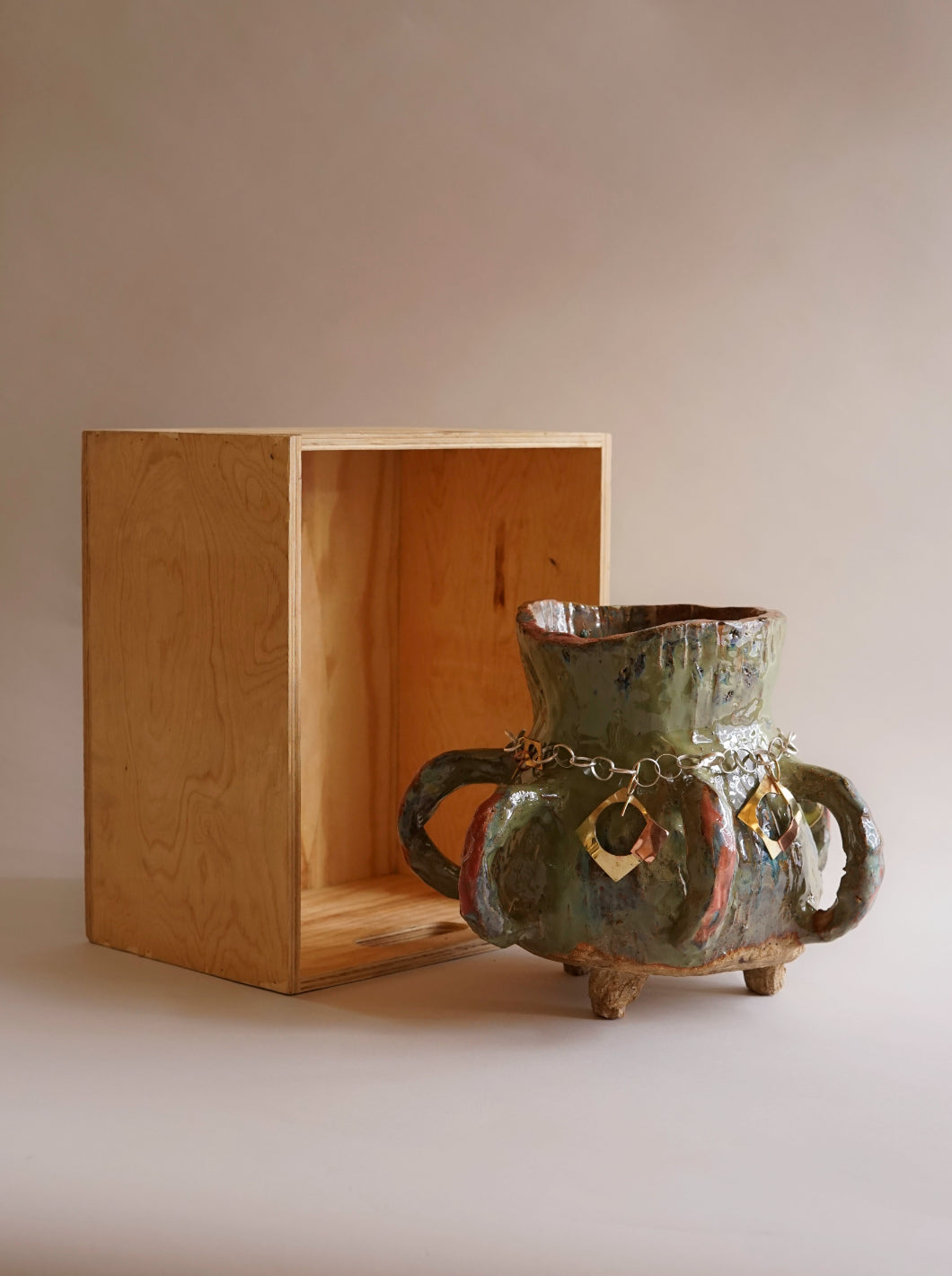  A stout ceramic pot with four circular handles, and three small feet. Around the neck of the vessel is a metal chain and pendant. The pot is next to a wooden box, turned on it's side.