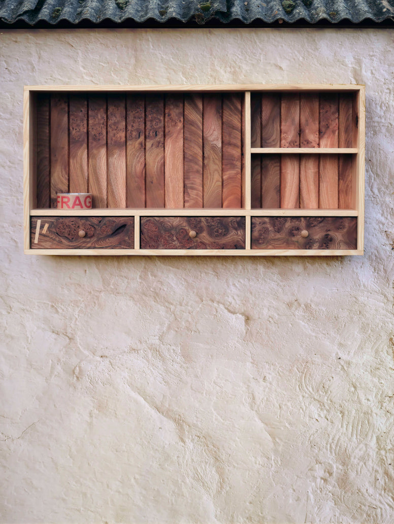  Tom Addy's Prìseil, is a wooden wall mounted storage shelf with three drawers. Photographed here outdoors, on a textured beige wall, with the weathered guttering visible at the very top of the image.