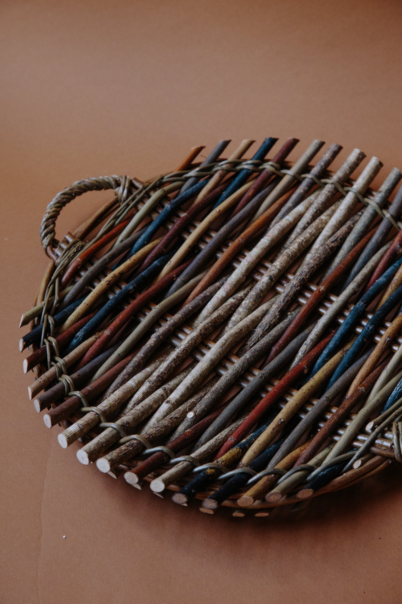 Circular willow platter woven from woods and willow with two small handles on either side. Photograph shot on a deep brown background.