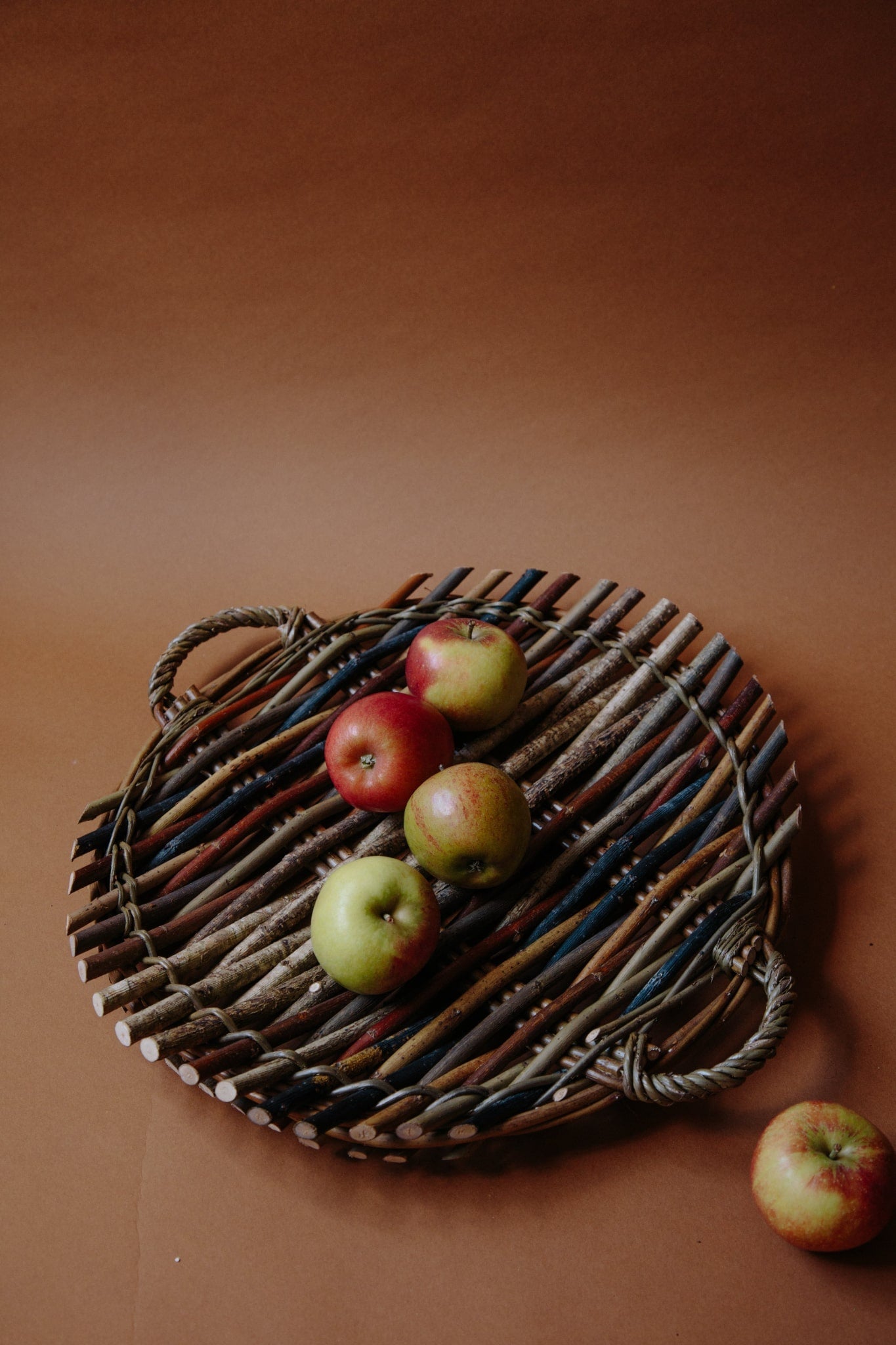 Circular willow platter woven from woods and willow with two small handles on either side. Platter holds four apples, with a fifth rolling out of frame. Photograph shot on a deep brown background.