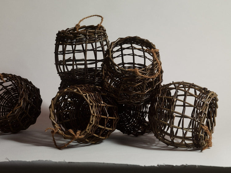 Small fruit and vegetable pots woven from willow with string handles. Six shown in frame, on a grey paper background. 