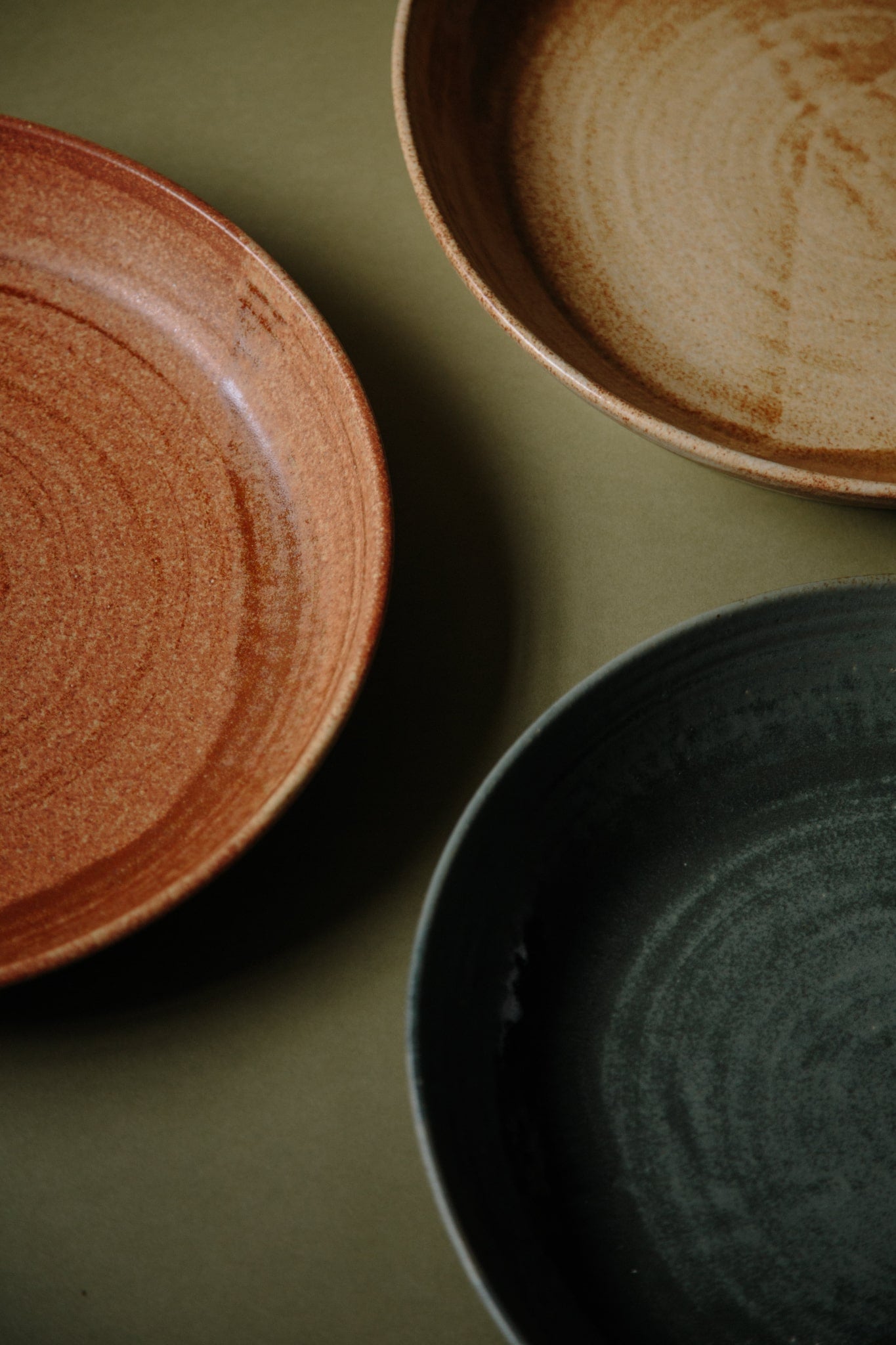 Group of three large ceramic platters in cool green, ochre yellow and red rust. Close up shot showing quarters or halves of each bowls on an olive green paper background.