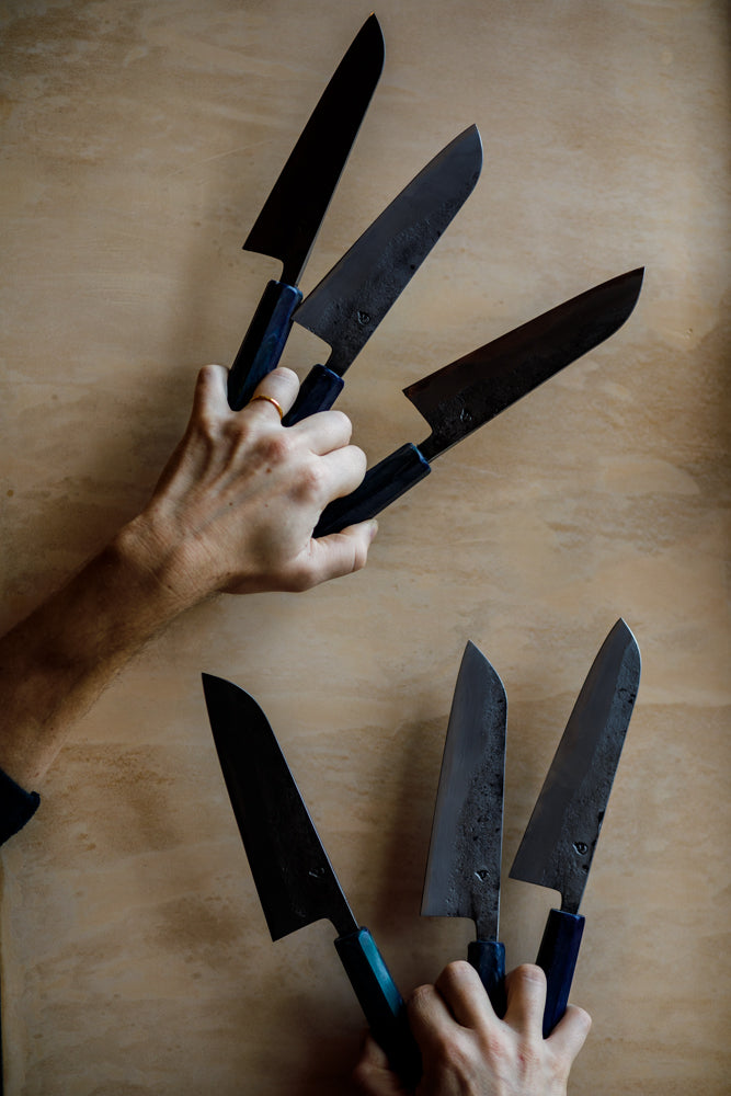 Six 6" chef knives by Clement Knives held between the forefingers against a wall at Bard Scotland.