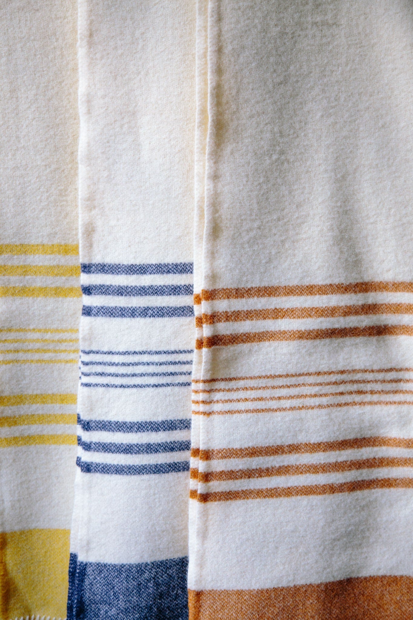 Three striped lambswool blankets in yellow, ochre and navy blue, hung vertically infront of eachother. The thick, weight of the blankets can be seen.