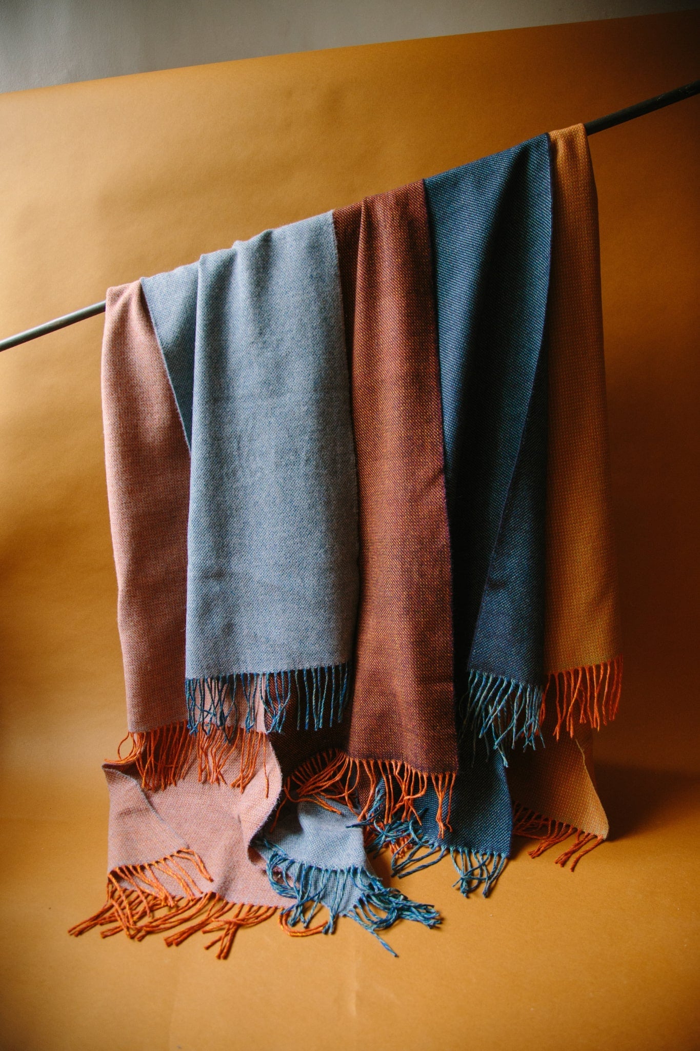 Group of five merino wool scarves hanging from a black curtain pole against a brown background. The scarves are in shades of reds and blues with tassels at the ends.