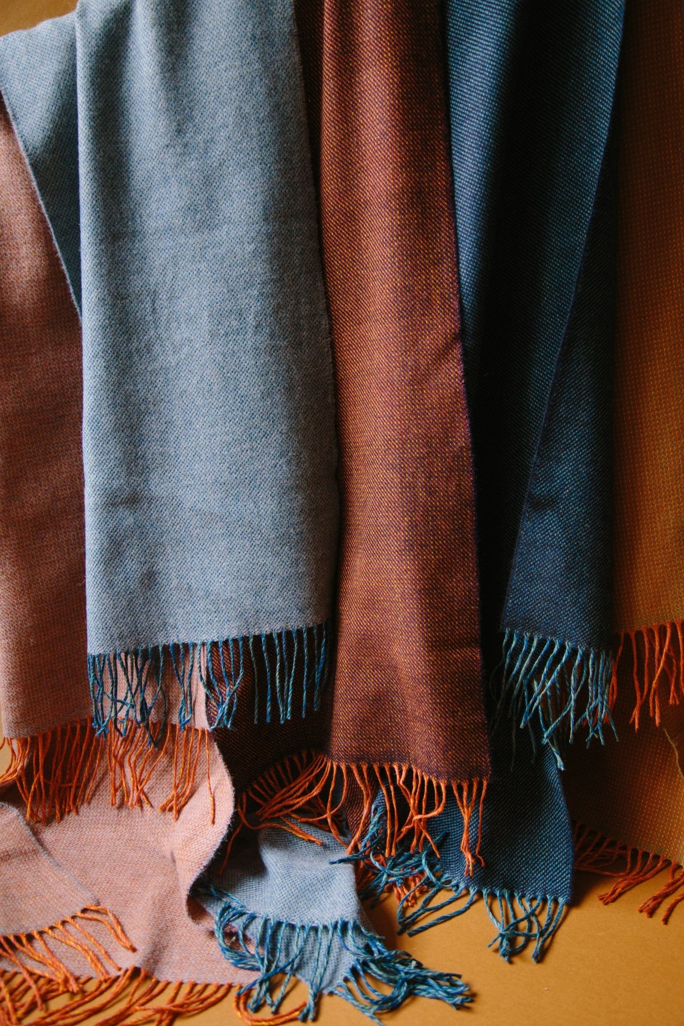 Close-up shot of five merino wool scarves, in shades of blue and red with tassels at each end. The scarves are hung from a black curtain pole against a brown background in natural light. 