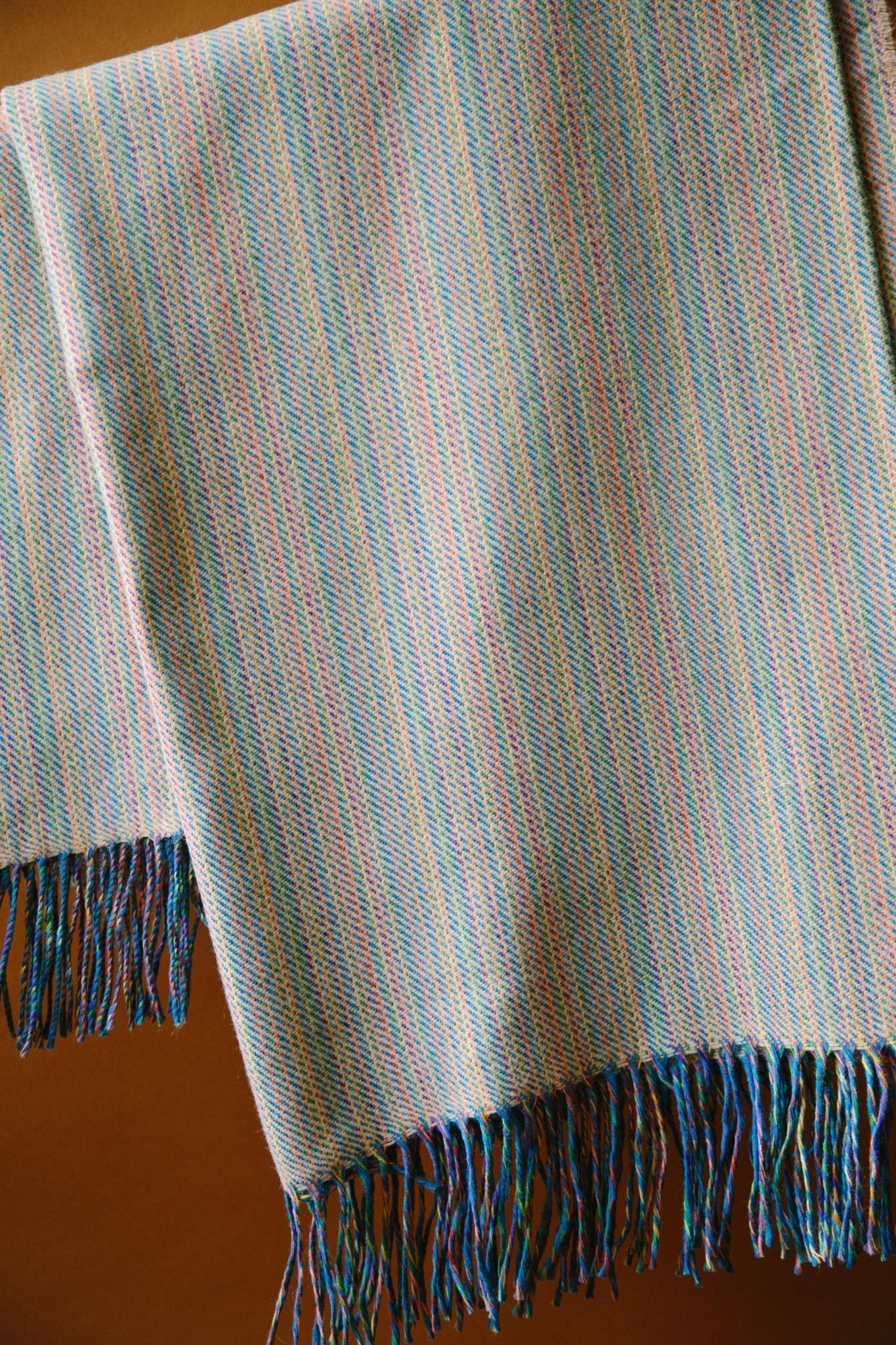 A merino wool throw hung from a black curtain pole. With multi coloured tones and ochre yellow with tassels at each end.