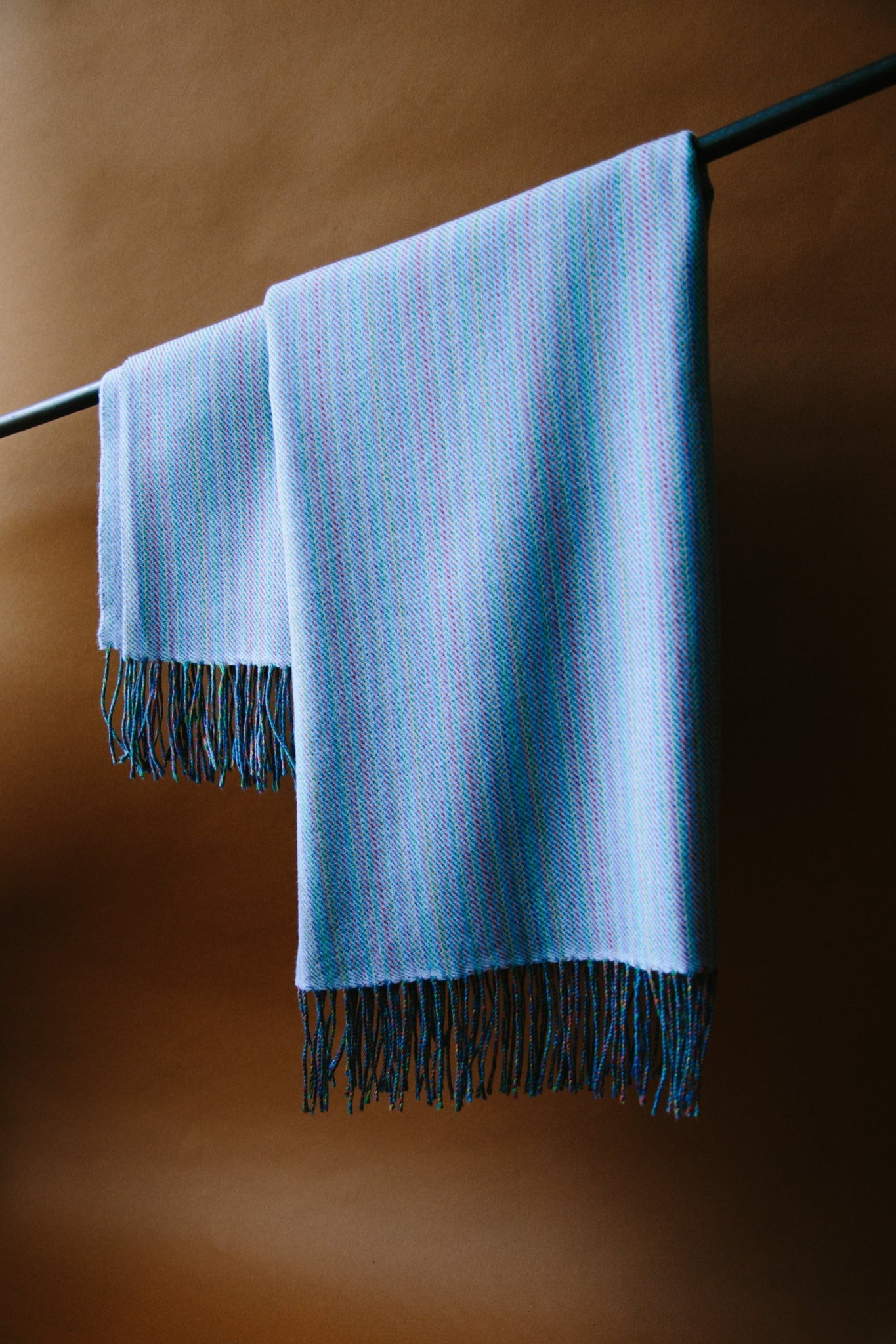 A sky blue merino wool throw hung from a black curtain pole. The throw has tassels at each end and is reflecting the natural light from a window on the left of the shot.