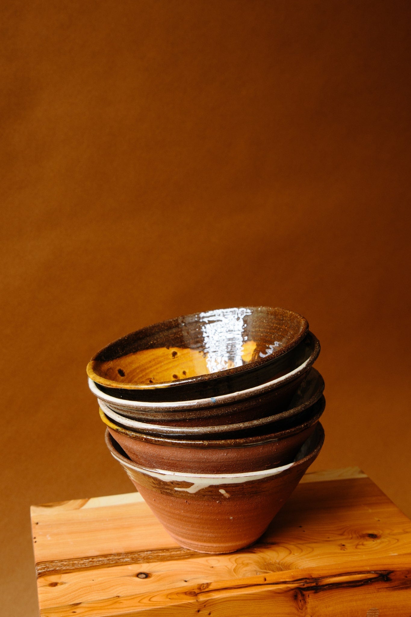 A pile of terracotta bowls against a deep brown paper backdrop. A beautifully uneven application of glaze along the bowls edge is viewable from this side angle.