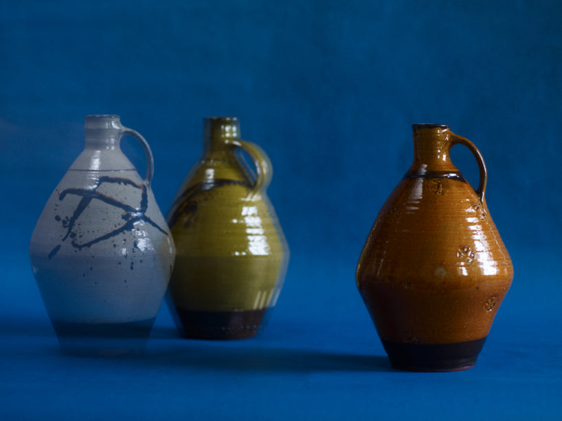 Three hand-thrown ceramic flagon jugs with primitive markings applied in a deep brown glaze. Made by Joshua Williams. Photographed alone on a blue paper background.