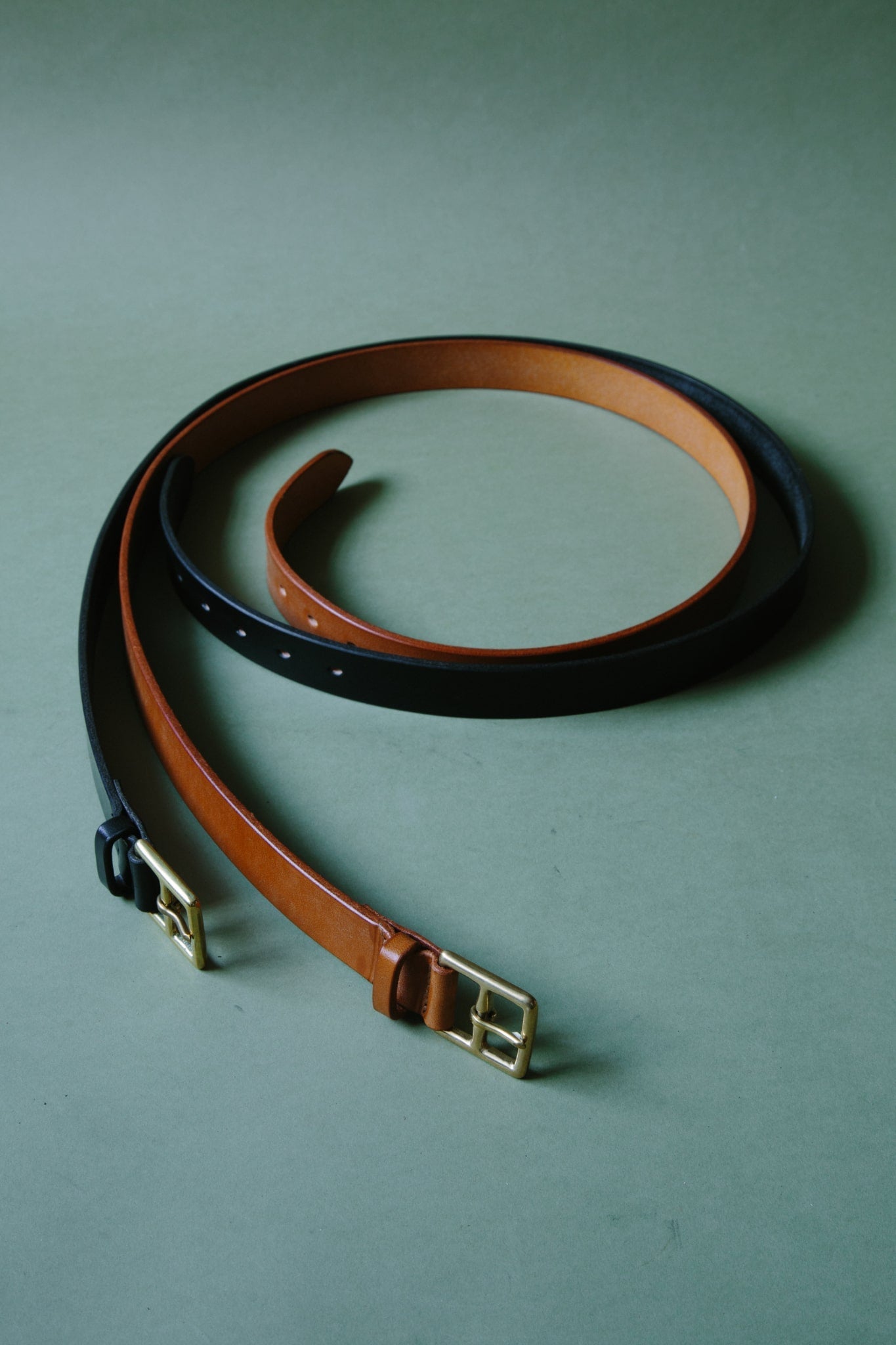 Two McRosite bridle leather belts, wrapped into circles, one tan and one black with metal buckles. Shot on a green background.