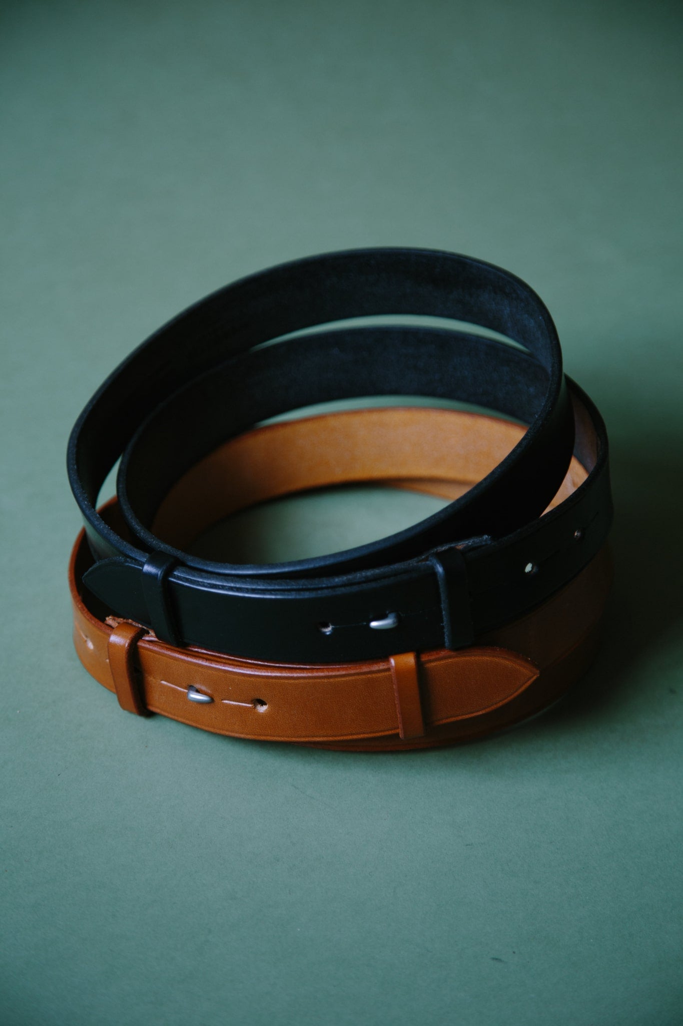 Two McRosite bridle leather belts, wrapped into circles, one tan and one black. Shot on a green background.