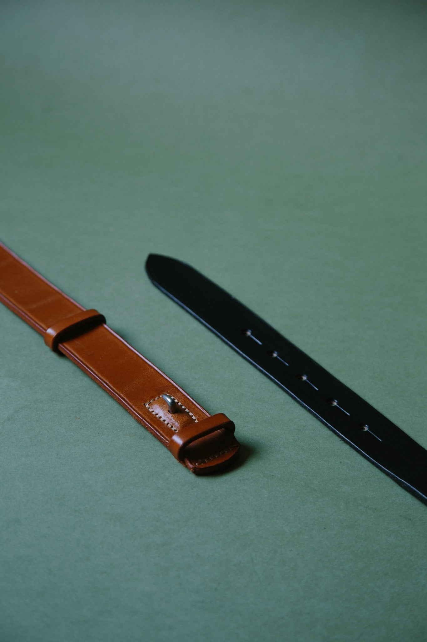 Two McRosite bridle leather belts, laid flat so the buckleless fastening can be seen. One tan and one black. Shot on green paper.