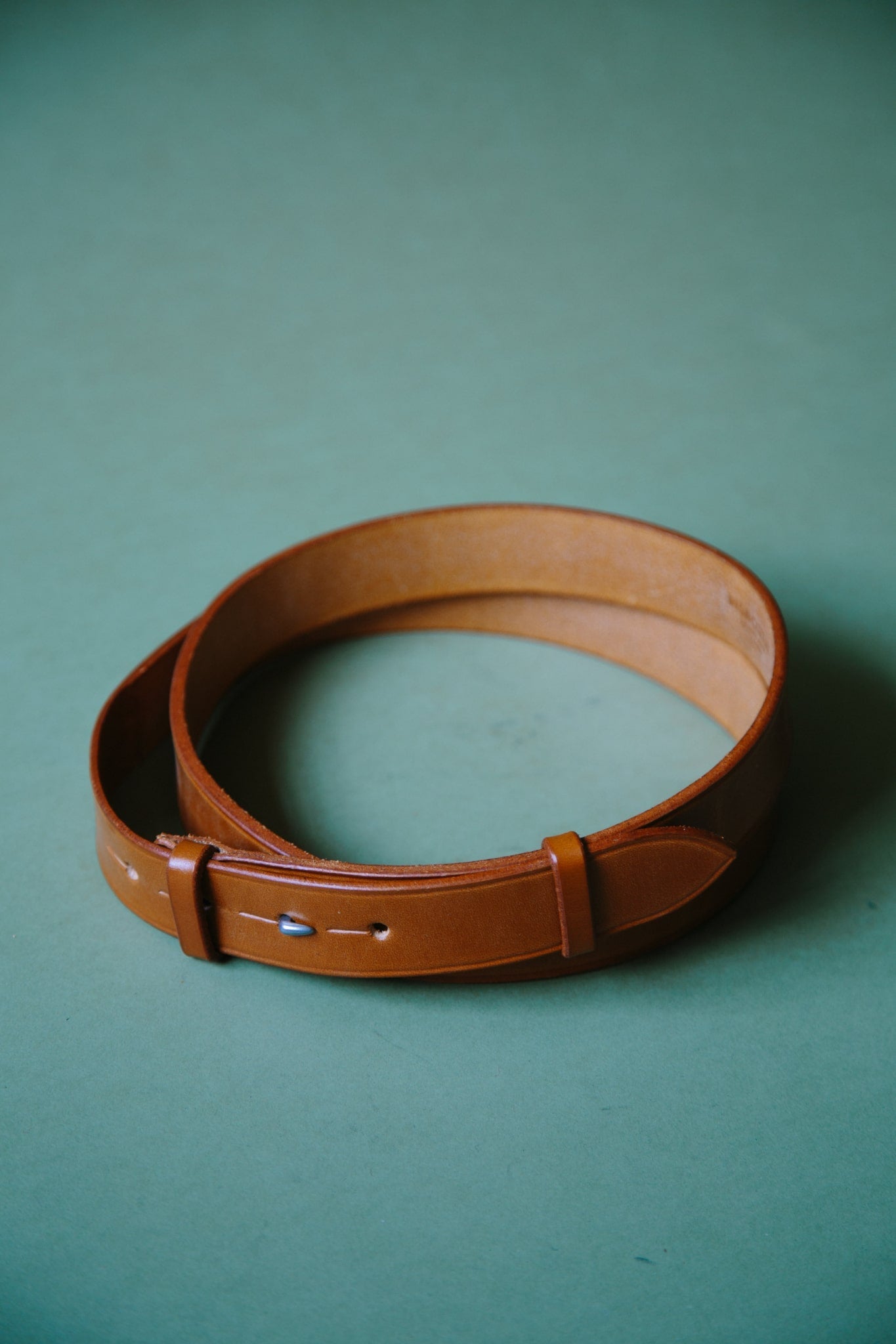 A tan bridle leather belt, wrapped into a circle so it fits within frame. The belt is buckleless and the hook and stud fastening can be seen.