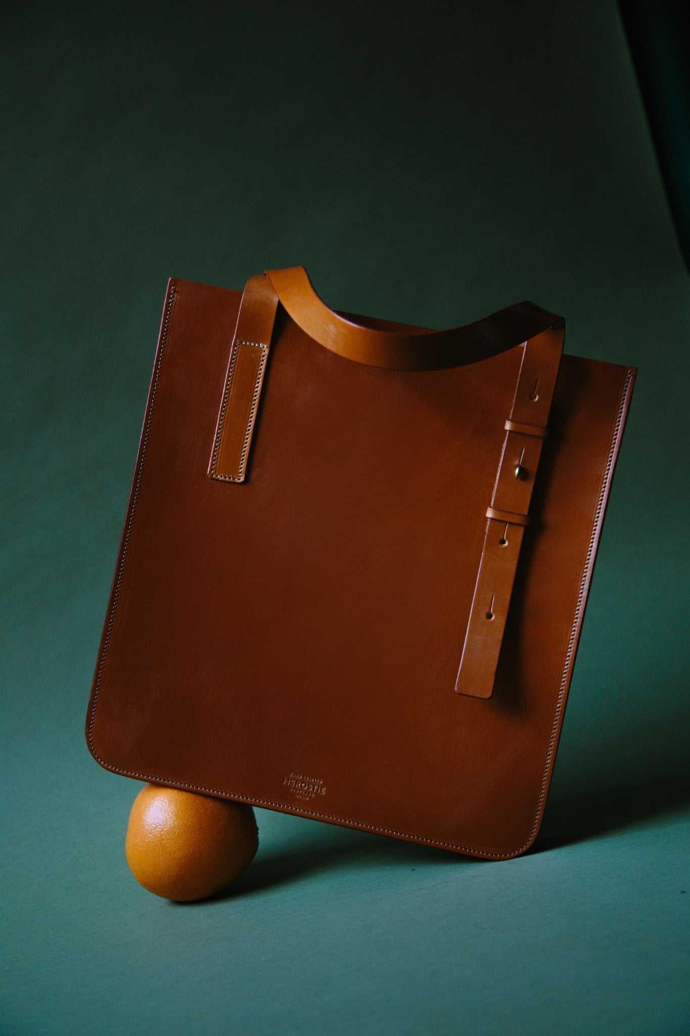 A large tan leather tote, balanced on an orange. The leather handle is illuminated in natural light.