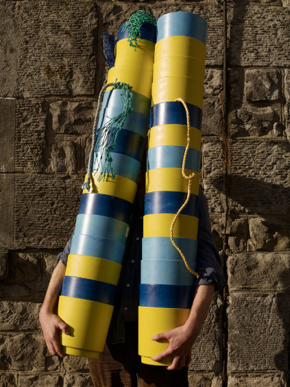 Two stacks of Burton recycled plastic plant pots, in yellow, blue and navy. Held by Hugo outside Bard in Leith.