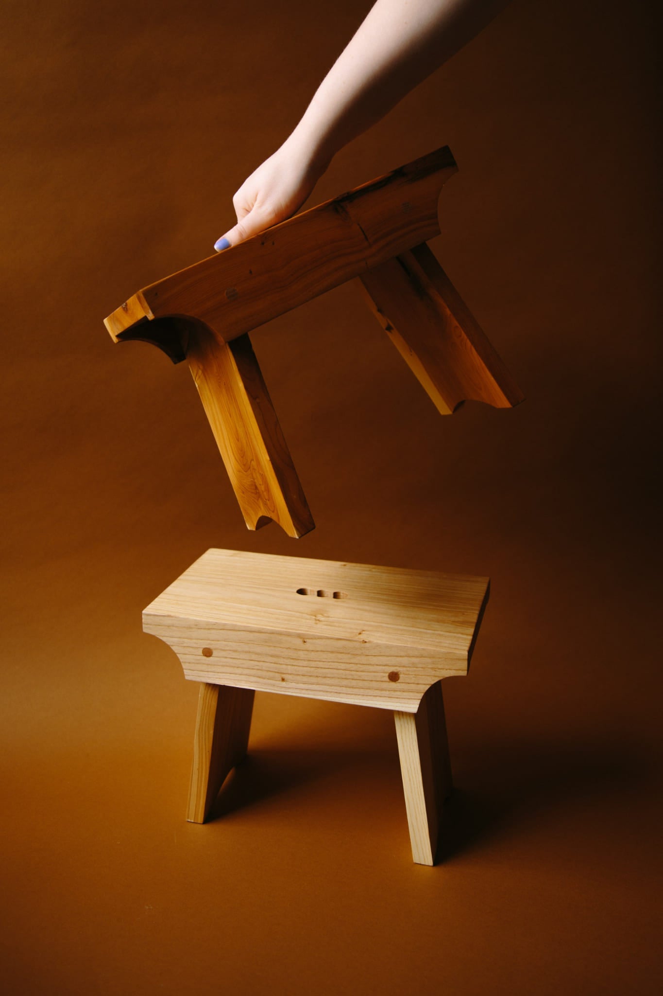 Two wooden shetland creepie stools in deep brown space. One being held by an arm coming in from the top of the frame. Held using the hand carved finger holes in the stools top.