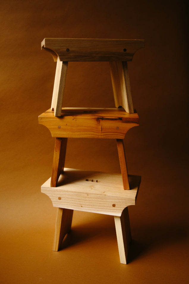 Three wooden shetland creepie stools stacked upon one another in a deep brown space.