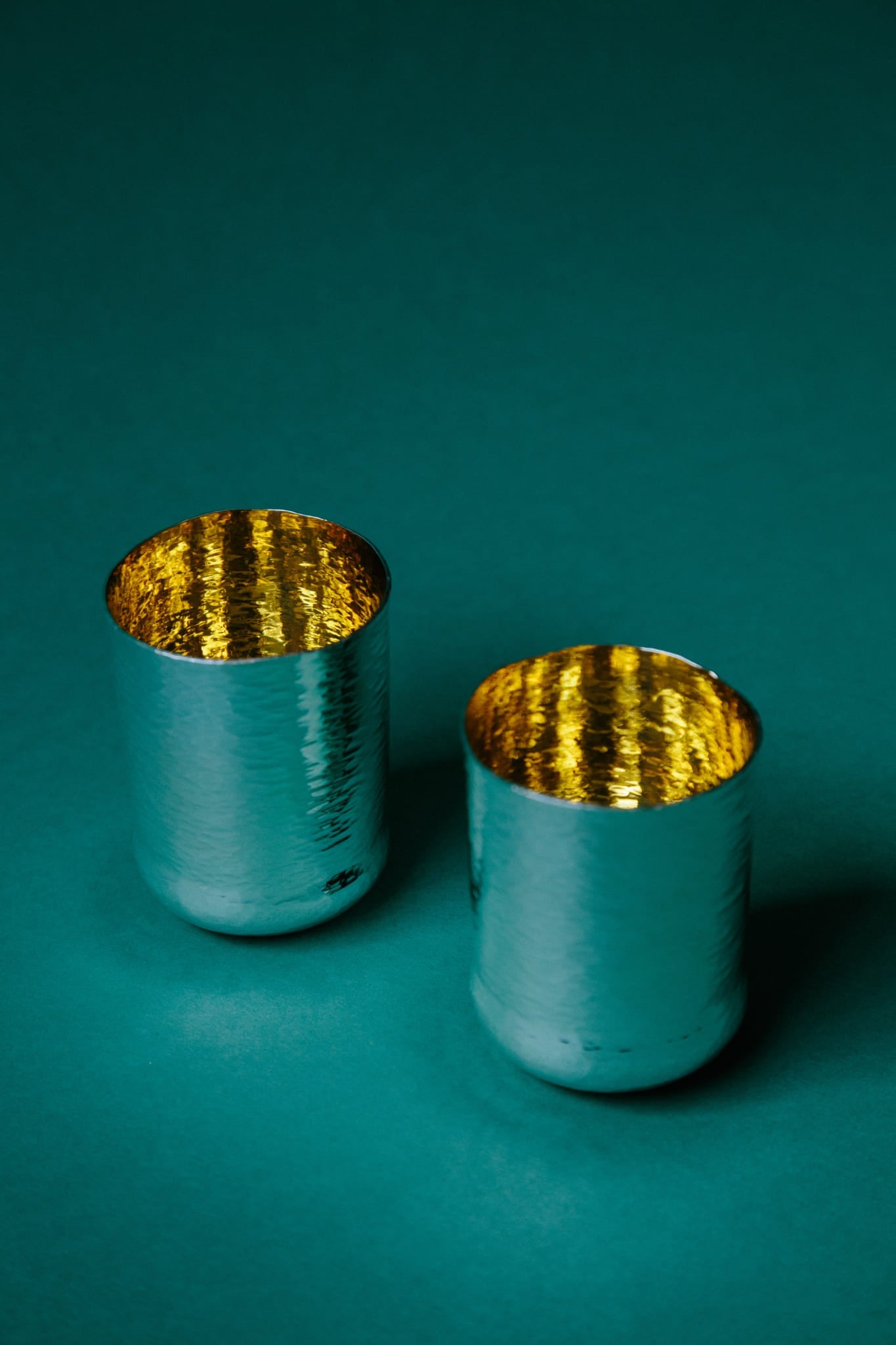 A pair of hand-raised sterling silver cups with gold-plated inner surfaces. Highly reflective inner surface, contrasting against a deep green paper backdrop. 