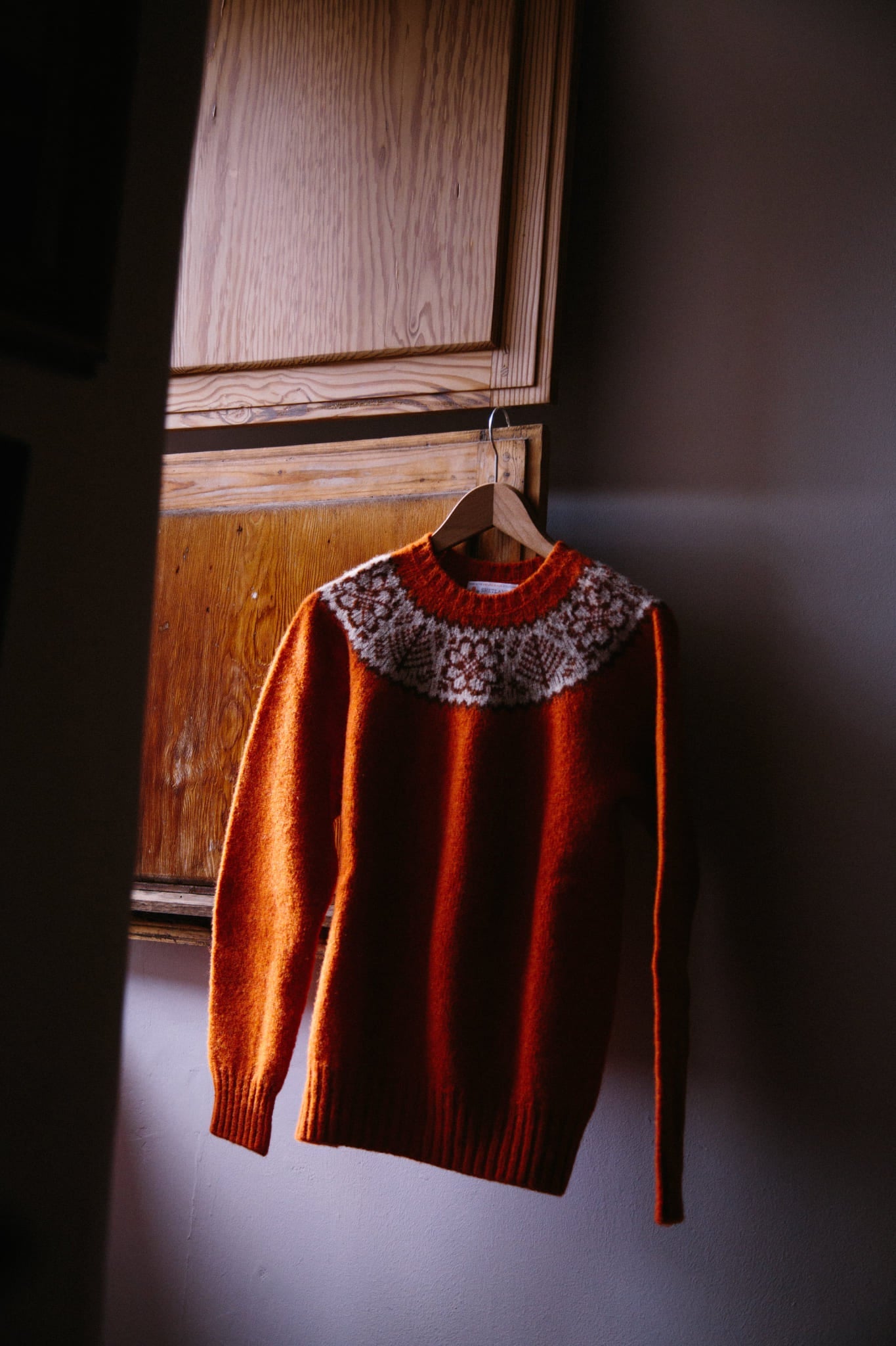 A Shetland woolen pullover jumper in Fair Isle Yoke, iron rust red. Hung on a wooden hanger from a window shutter.