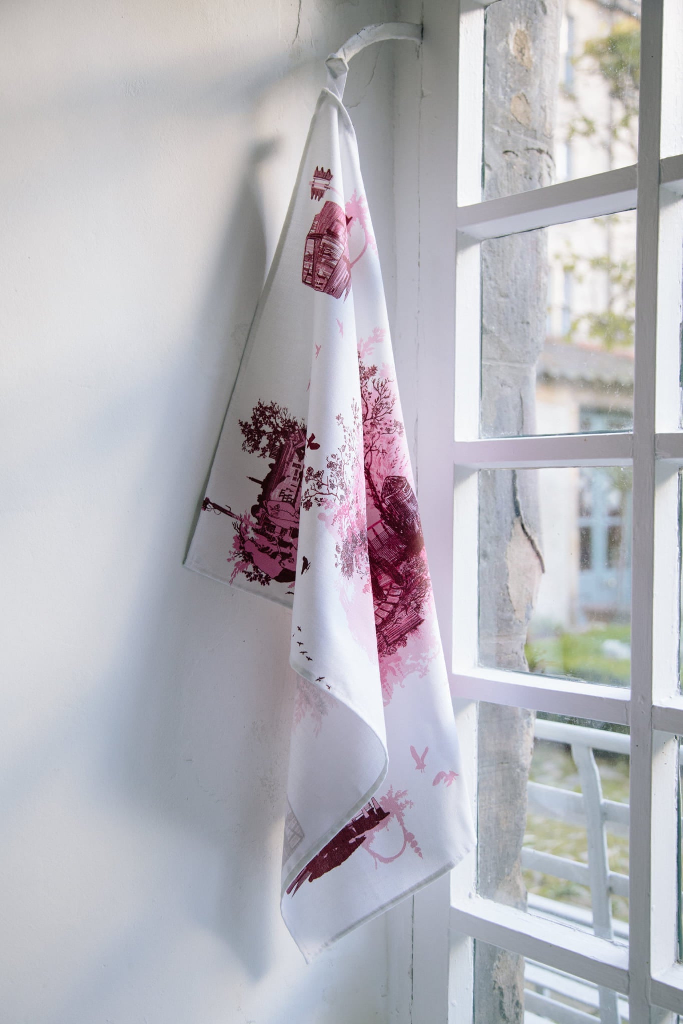A cotton tea towel hung from a window hook. Printed with a pink toile design. The design shows the gritter side of Edinburgh.