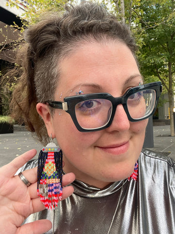 Sara, smiling at the camera with her hand behind an earring - a beaded UFO with a beam that is multi-coloured a la designer Pucci. Sara is a fat white-skinned woman with shaved sides of her head, and messy wavy brown hair with lots of greys peeking through
