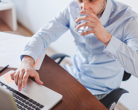 Hombre bebiendo un vaso de agua mientras trabaja en su computadora.