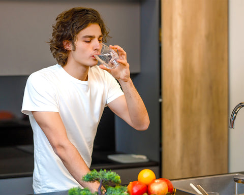 Hombre tomando un vaso de agua en su cocina en la que tiene vegetales.