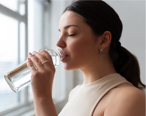 Mujer bebiendo agua de su botella y al lado de una ventana