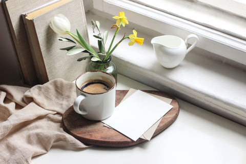 books, spring flowers in a vase, mug of coffee and card on a wooden disc, cloth napkin and coffee creamer dish all by a window