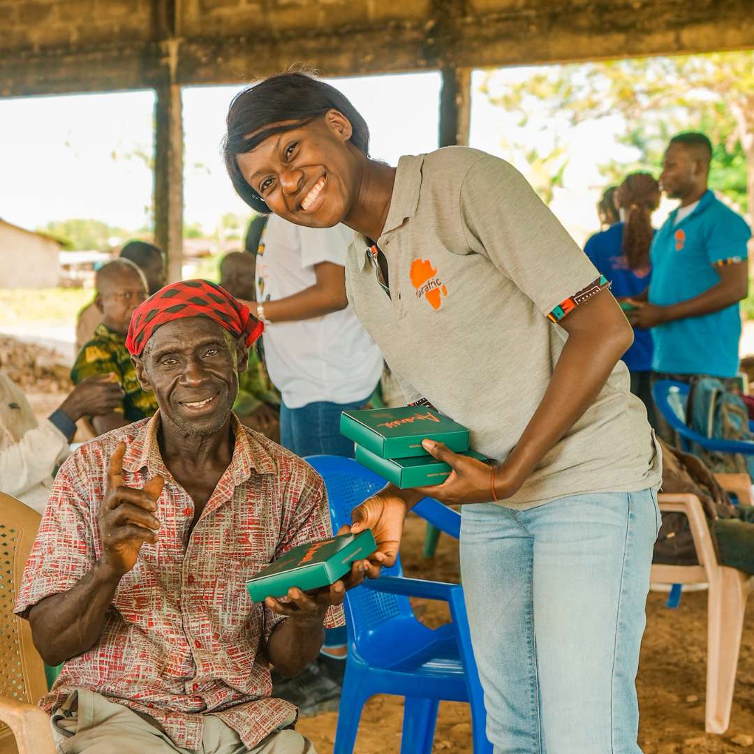 A smiling woman handing a box to an elderly man wearing a red headscarf, with others in the background.