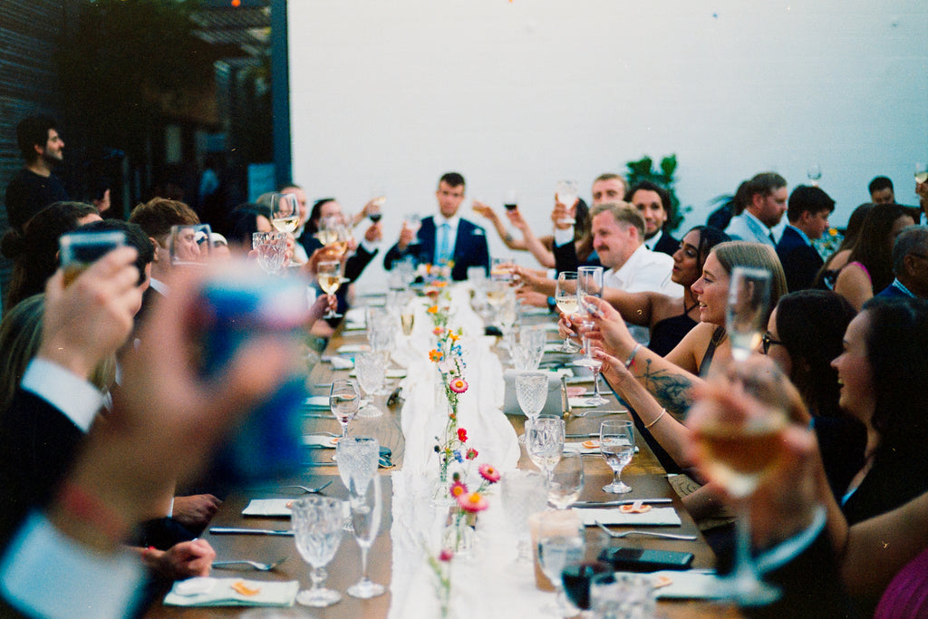 Guests doing cheers at a long table with colourful flower centrepieces.