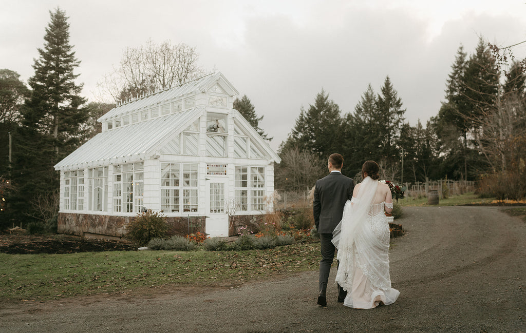 bride and groom walking on a gravel path towards a white glass green house