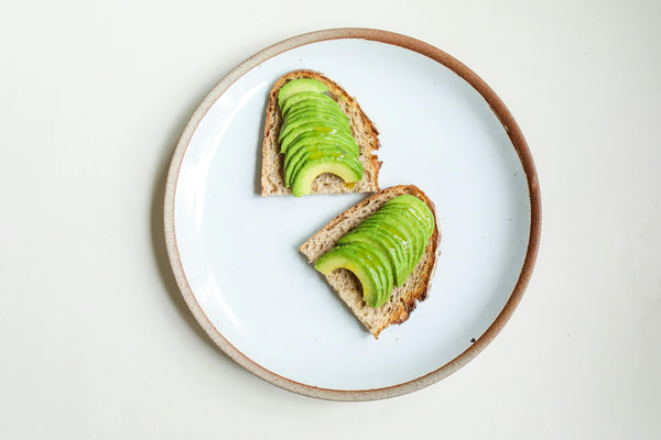 Fresh avocado slices neatly arranged on multigrain bread, served on a minimalist white plate with a brown rim
