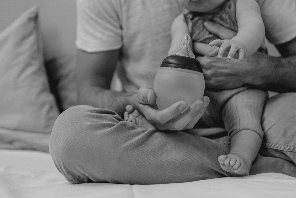 a father holds his baby's bottle, they are seated on a bed