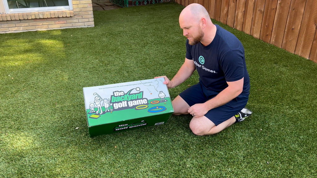 Man crouched in backyard with game box talking about The Backyard Golf Game review.