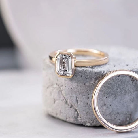 an emerald cut diamond is bezel set on a narrow yellow gold ring