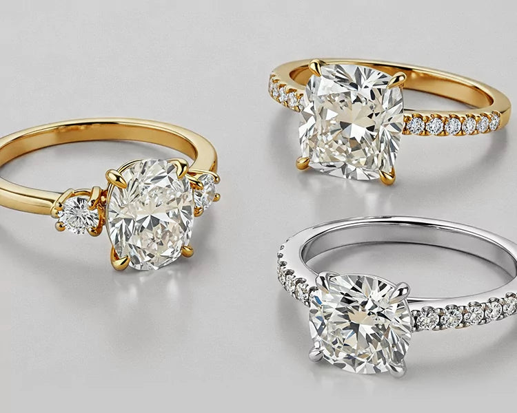 Two are solitaire rings with a cushion-cut diamond center stone, set in yellow gold and white gold respectively, and both feature a pave diamond band. The third ring is a three-stone setting in yellow gold, with a larger cushion-cut diamond in the center and two smaller round diamonds flanking it.