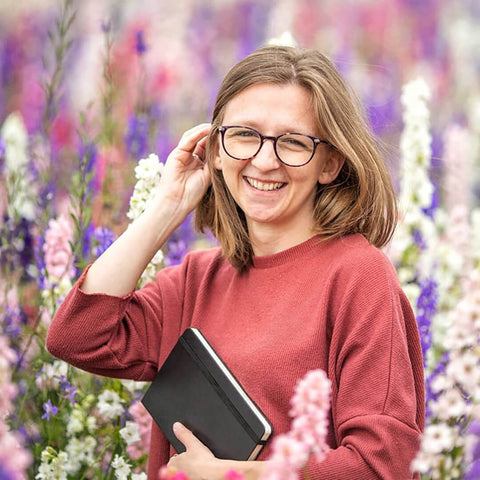 Headshot of Jess wearing red jumper and carrying a black notebook, stood in a field of pink, purple and white wildflowers