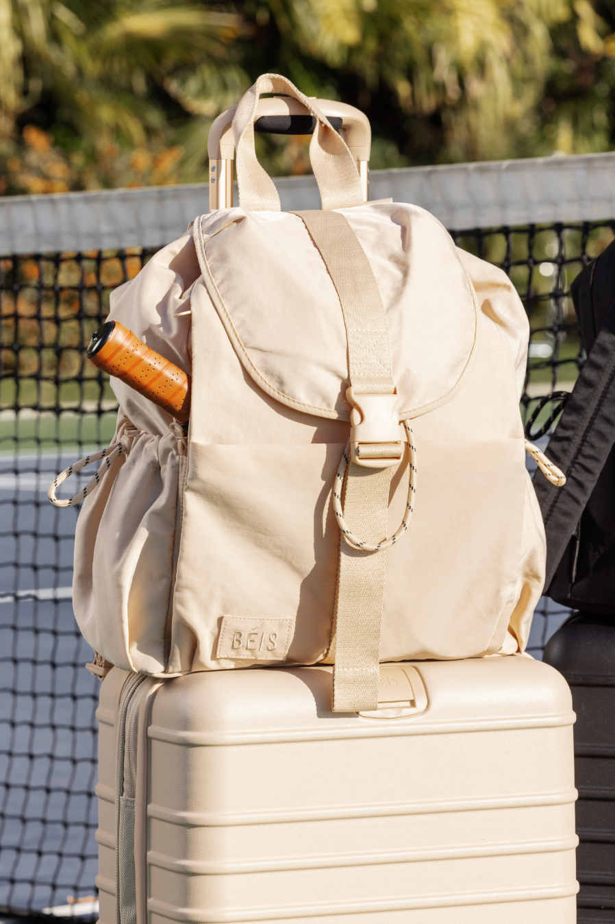 Beige beis backpack with a tennis racquet inside On top of a beige suitcase on a tennis court