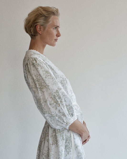 Side view of a woman leaning back wearing a long-sleeve dress. Dress is floral cotton with puff sleeves and eyelet trim.