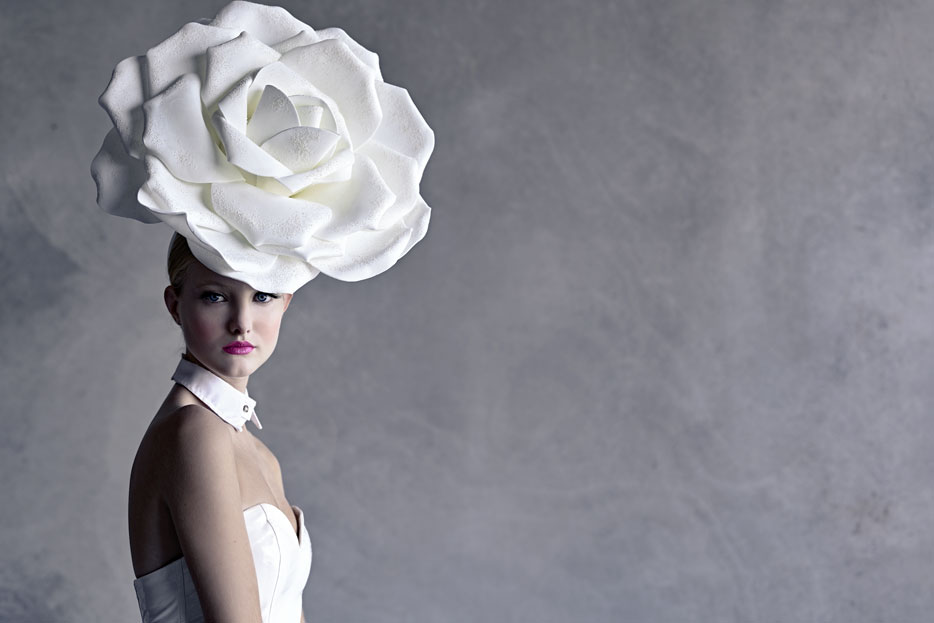 Woman in white dress with a large white rose on her head against a gray background 