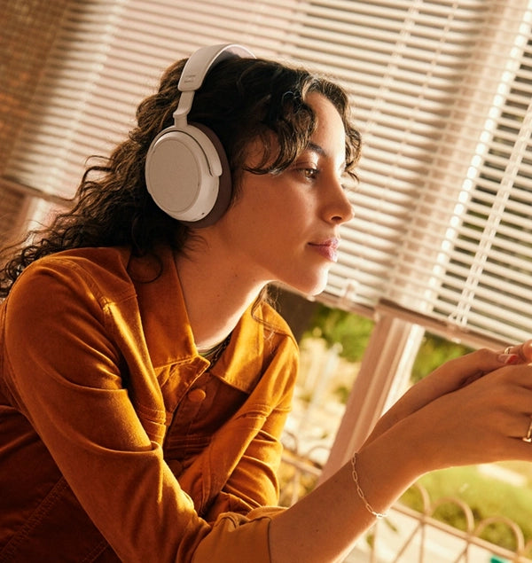 A woman in a sepia t-shirt stands in front of a window, wearing white SENNHEISER ACCENTUM PLUS headphones.