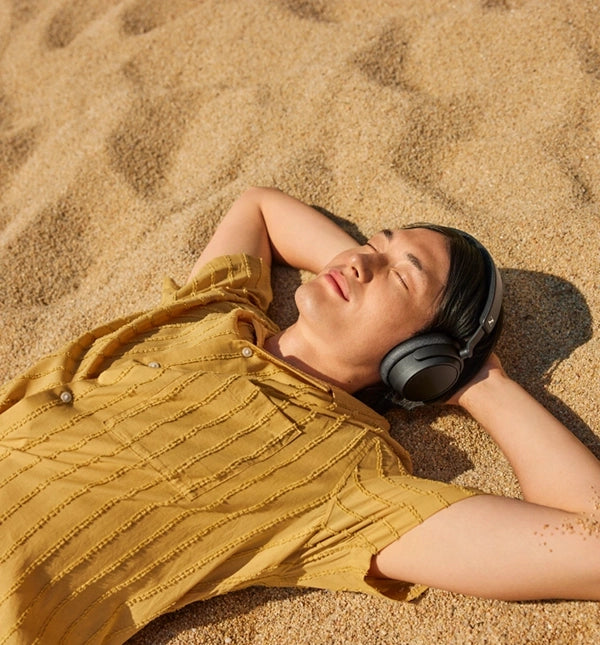 A man resting on the sand and wearing black SENNHEISER ACCENTUM PLUS headphones.