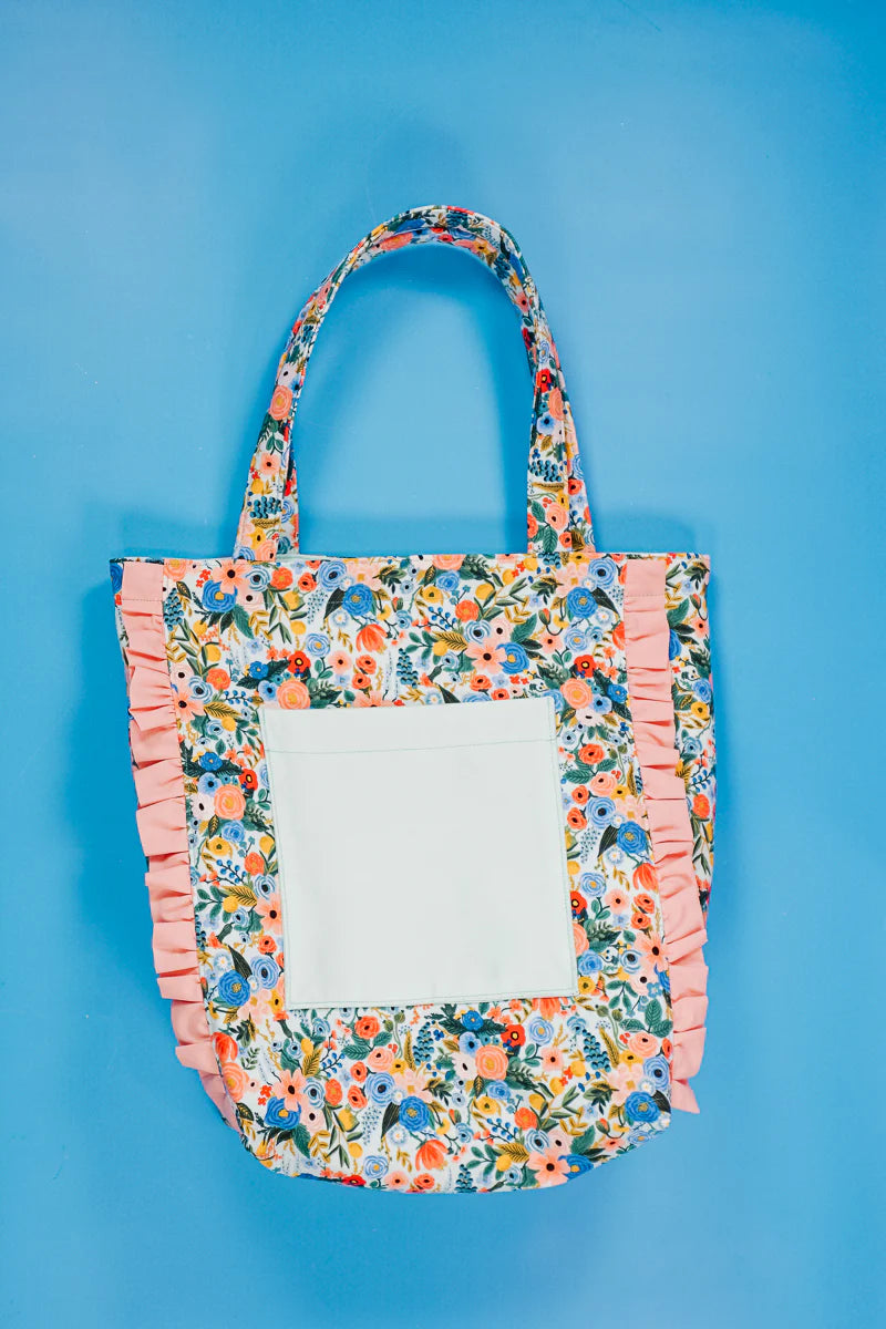 Floral tote bag with pink ruffled edges and a front pocket on a blue background.