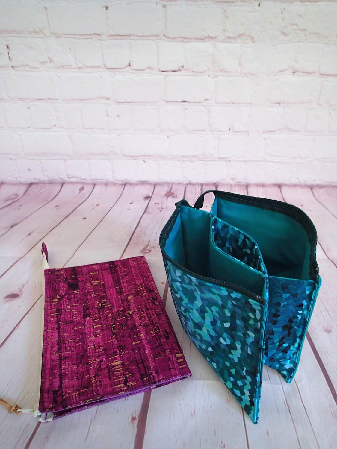 A pink zippered pouch and a blue patterned organizer on a wooden floor with a white brick wall background.